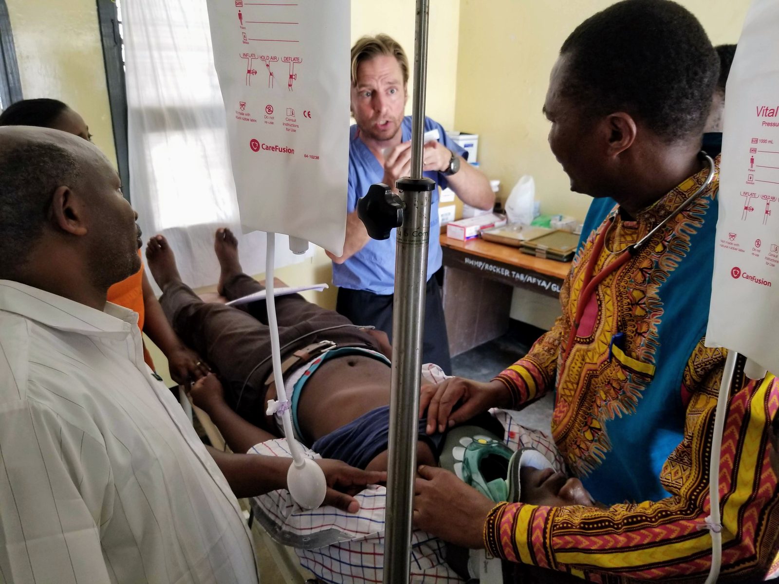 Ben Belknap in blue scrubs teaching Tanzanian doctors gathered around a trauma patient in a cervical collar, with pressure bags of IV fluid hanging overhead