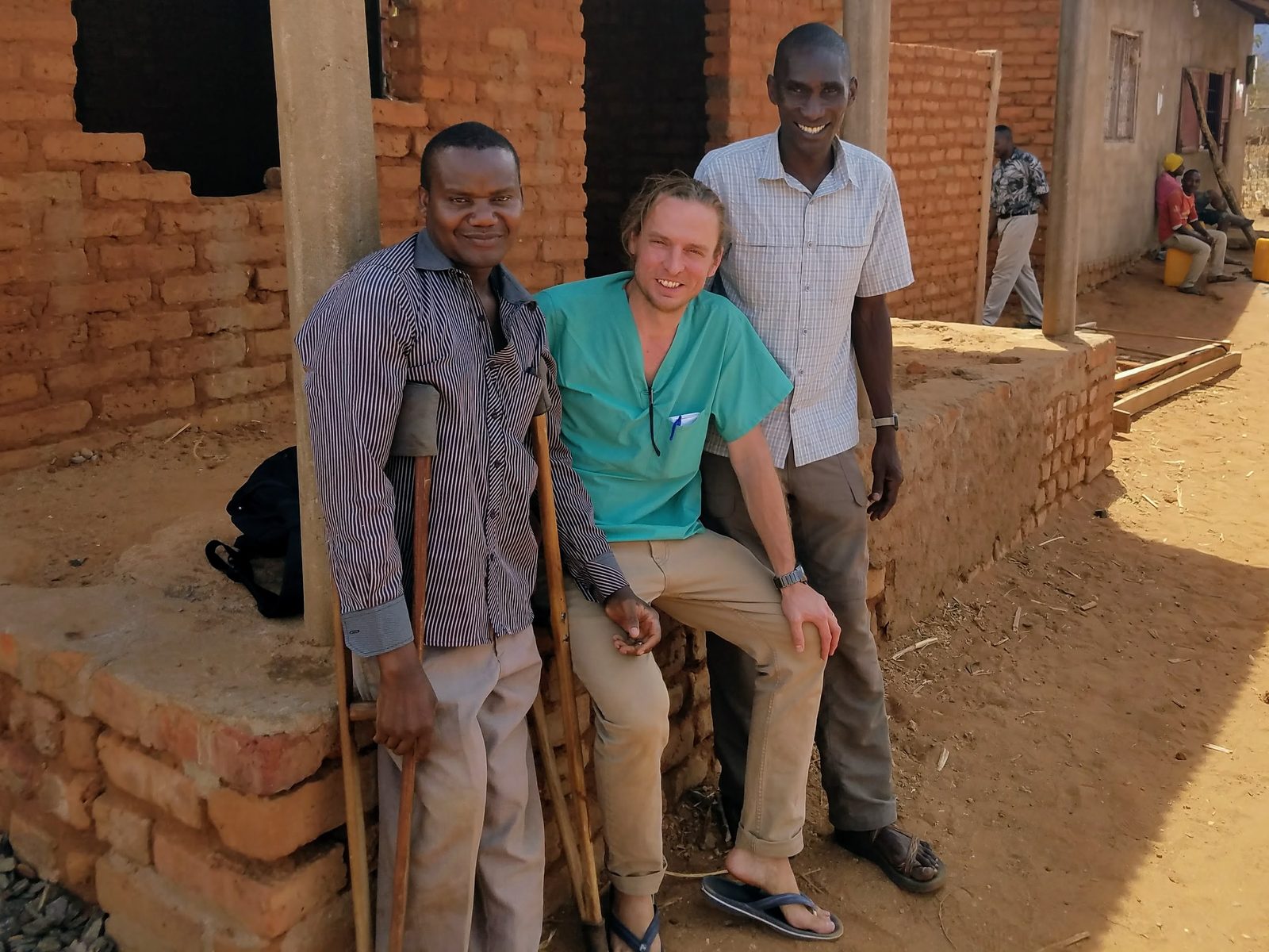 Ben Belknap in scrubs, seated between Laithon Mvili and a friend outside a mud-brick building in Kingiti