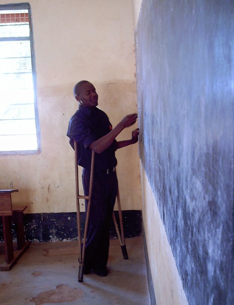 Laithon Mvili, standing on crutches, writing on the chalkboard in a Kingiti Primary School classroom