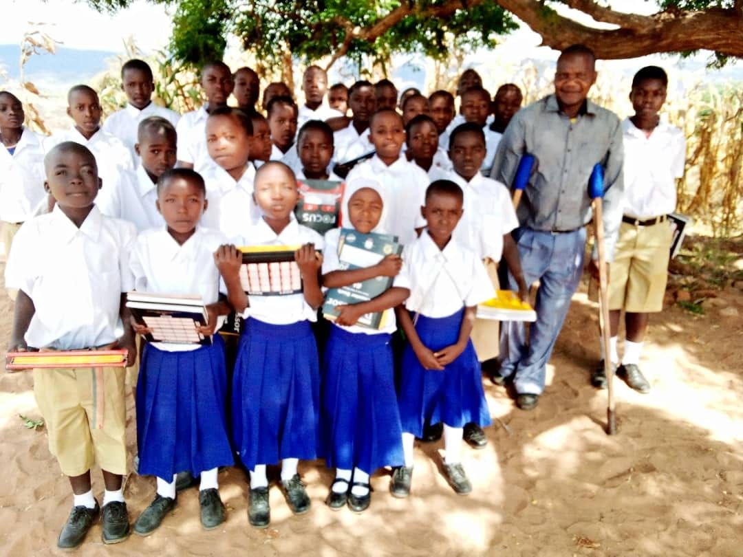 Secondary school scholarship students in white shirts and blue skirts holding new school supplies under a large tree
